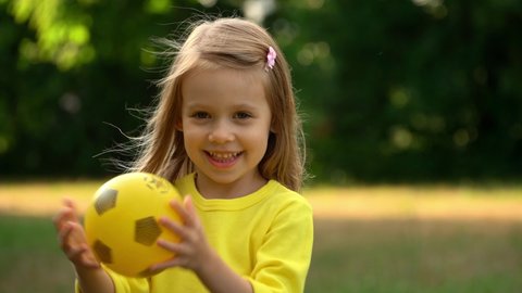 Little girl with yellow ball on green lawn in summer park at sunset. Happy child throws and catches soccer ball, smiles and laughs. Football and sport concept.
