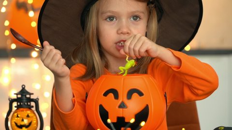 Little girl in witch costume fun brews potion from spiders, snakes, frogs in pumpkin pot, stirs with spoon and tastes. Halloween.