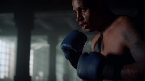 Tired athlete training boxing with punching bag in dark gym. African american man working out blows on sports bag. Active sportsman in boxing gloves cardio workout. Closeup professional boxer exercise
