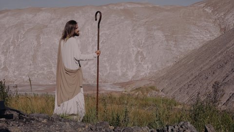 Jesus Christ with a staff in robe stands on the mountain and looks into the distance. There are bruises on his face, wounds on his hands.