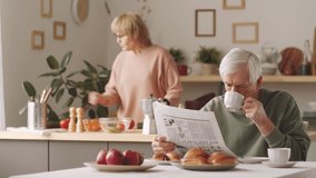 Elderly Caucasian man drinking coffee and reading newspaper at kitchen table while his wife cooking meal in background - Powered by Shutterstock - Get 15% off with code: PIKWIZARD15