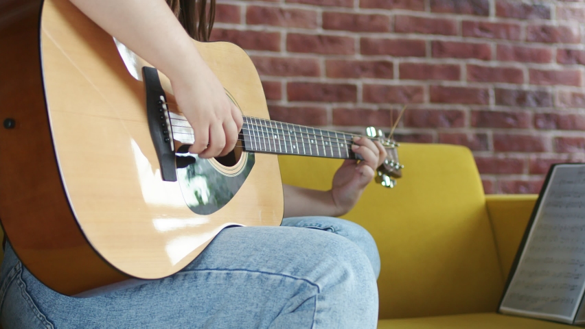 Woman is sitting on sofa and playing an acoustic guitar strumming strings. Woman is engaged in musical hobby her room