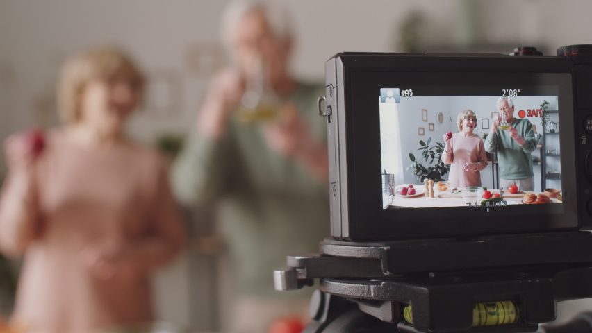 Close up selective focus shot of display of digital camera recording elderly couple giving culinary class in kitchen
