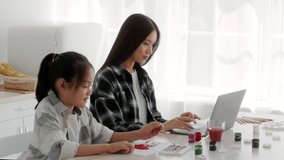 Asian Freelancer Mother Working Online On Laptop While Her Daughter Drawing With Watercolor Sitting Together In Kitchen At Home. Remote Job And Freelance Concept. Slowmo