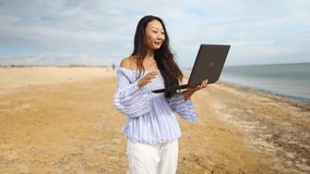 Asian business woman greeting during videocall on laptop sitting on the beach. Freelancer and blogger participates in Videoconference