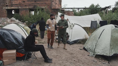 Tracking shot of social worker and police officer in military uniform with clipboard walking through tents of refugee camp where diverse immigrants and homeless people living
