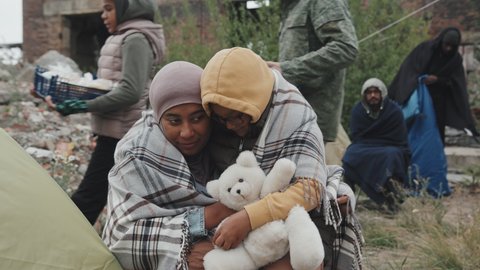 Medium shot of African-American mother and her 8-year-old daughter in warm clothing sitting outdoors under blanket embracing living at refugee camp with other immigrants