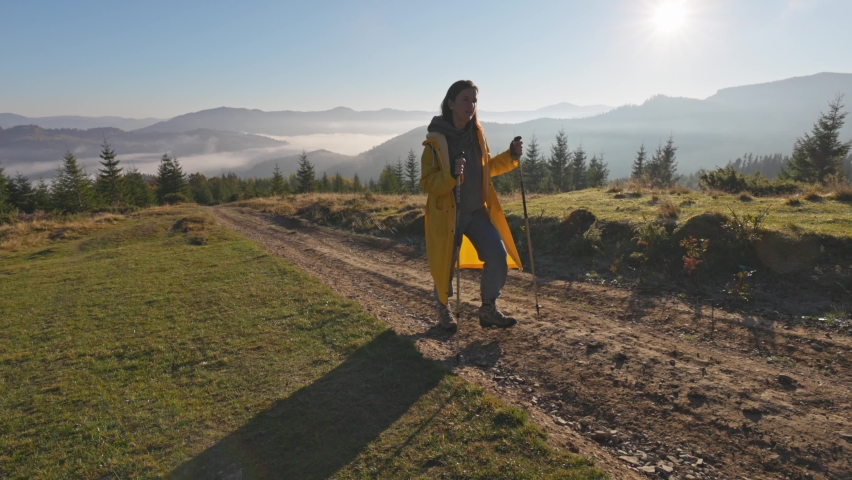 Woman in yellow raincoat walks along the road uphill. Morning sun and foggy mountains on the background. Travel hiking in nature concept, slow motion