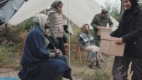 Stab shot of homeless Arab man and African-American girl living with other refugees in tents accepting donation boxes from social workers and volunteers