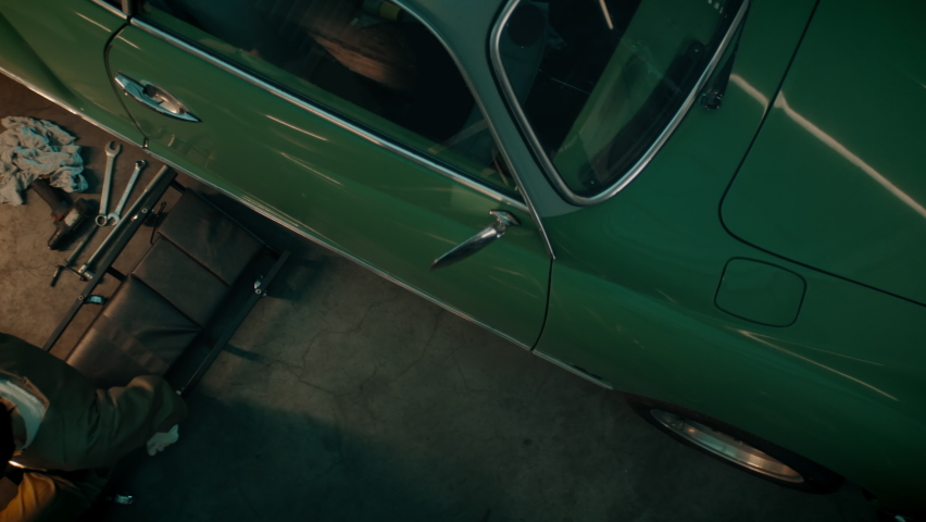 OVERHEAD High angle shot of Caucasian female mechanic repairing a vintage old car in a workshop, working under car bottom. Shot with 2x anamorphic lens Royalty-Free Stock Footage #1080621605