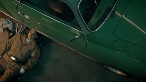 OVERHEAD High angle shot of Caucasian female mechanic repairing a vintage old car in a workshop, working under car bottom. Shot with 2x anamorphic lens
