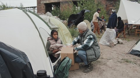 Stab shot of social workers carrying cardboard donation boxes giving warm clothes to diverse men, women and children living in tents at refugee camp