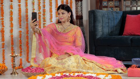 An elegantly dressed female wishing her friends on Deepavali on a video call. A young woman in ethnic attire sitting near a well-lit brass Diyas and a traditional Rangoli - Diwali decorations
