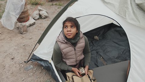 From-above shot of unhappy African-American girl in warm clothes sitting inside tent at refugee camp with tin of food in hands looking out at sky in hope