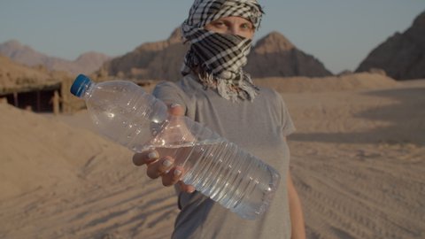 Woman in checkered keffiyeh holding bottle of water standing in the desert. Caucasian female tourist with plastic water bottle. Importance of drinking water.