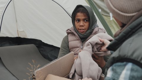 Medium close-up of female social worker with cardboard donation box giving warm clothes to African-American girl sitting in tent at refugee camp