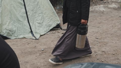 Tilting-up tracking shot of Muslim woman wearing black headscarf walking along poor tent city where diverse homeless and refugee people living