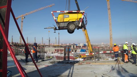 OMSK, RUSSIA - FEBRUARY 04 2021: Truck crane lifts up yellow machine under workers control at snowy construction site of sports arena against blue sky on February 04 in Omsk