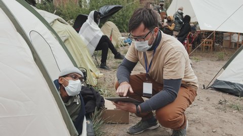 Tracking shot of social worker in face mask taking notes on digital tablet while talking to male immigrant sitting in tent at poor refugee camp