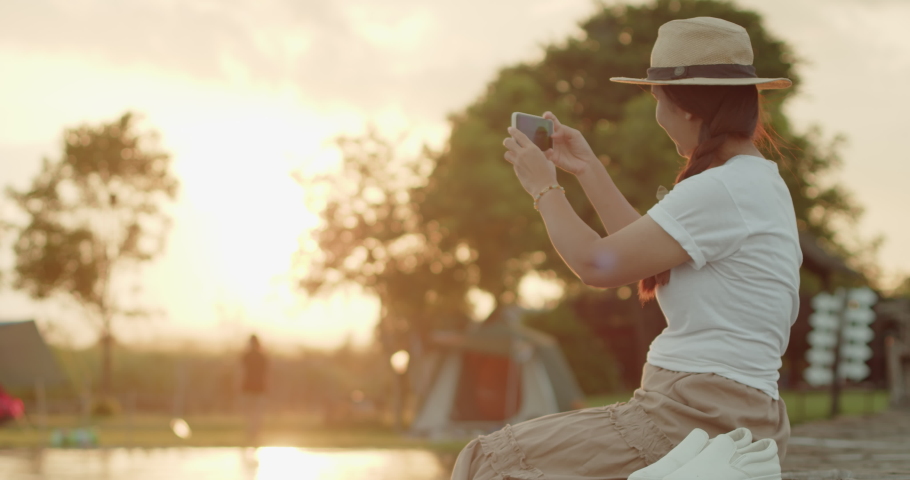 A female tourist in a brown hat, white T-shirt and brown skirt sits on a wooden bridge by the lake, using a mobile phone to take a beautiful view at sunset, an orange light reflecting on water surface