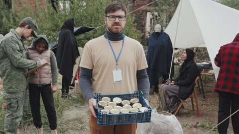 Medium portrait of male volunteer holding plastic crate with tinned food looking at camera standing at refugee camp among diverse immigrants living there