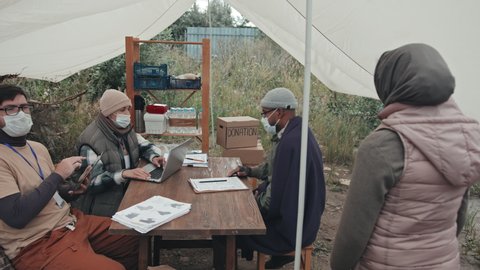 Medium shot of couple of social workers in face masks with laptop and digital tablet sitting under tent in poor refugee camp talking to immigrants, sitting down at their table one by one