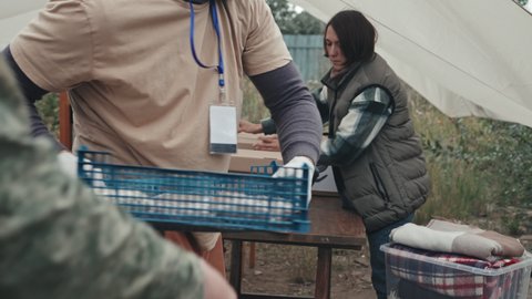 Midsection shot of police officer in military uniform helping social workers to prepare donation boxes with food and clothes for refugees living in poor tent city