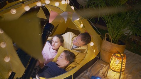 Happy father with son and daughter uses tablet spending time together in spacious play tent with fairy lights in cottage yard in summer evening