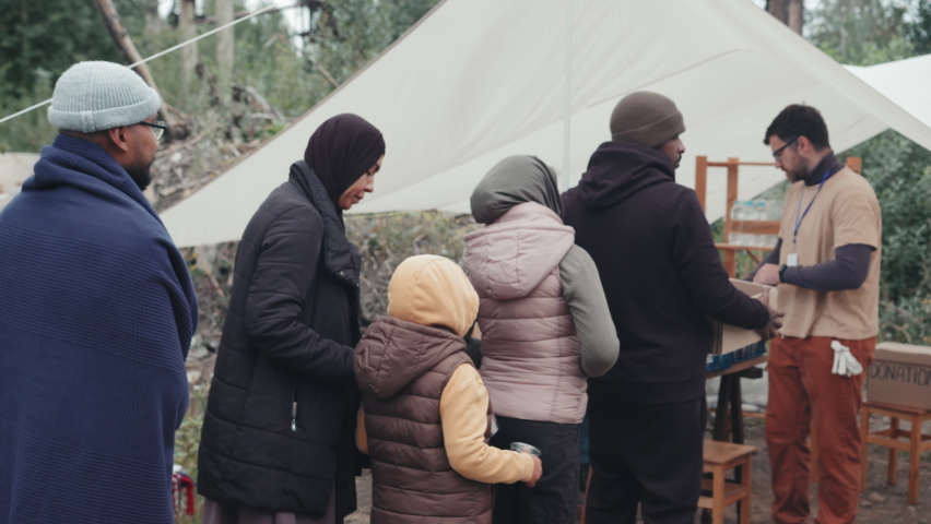 Tracking shot of diverse homeless people and refugees living at tent city standing in queue for getting boxes with food, water and clothes from social workers Royalty-Free Stock Footage #1080631283
