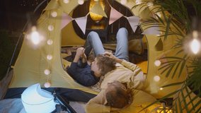Daddy with little son looks at stellar map on mobile phone spending time together in play tent with fairy lights in cottage yard in summer evening