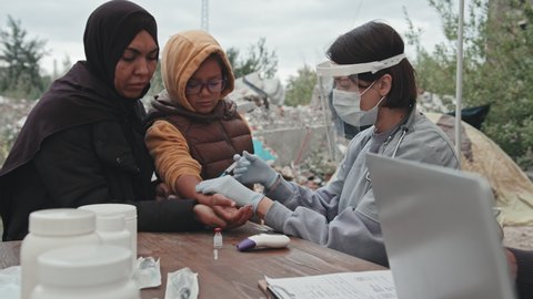 Waist-up shot of little refugee girl standing next to her mother getting vaccinated by female medical worker wearing face shield at tent city