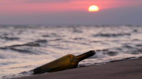 Sealed bottle with paper message washed ashore and lie on sand. Sunset sky and wavy sea seen blurred on background. Windy weather, small waves splash on beach