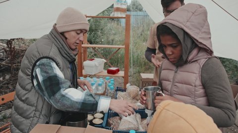 Medium slowmo shot of couple of social workers providing hot drinks and food to immigrants living in poor conditions at refugee camp