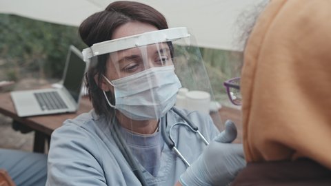 Medium close-up of female medical worker in protective clothing, face shield and mask examining child at refugee camp