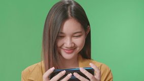 Close Up Of Smiling Asian Woman Looking At The Phone Screen And Holding It In Her Hands While Standing On Green Screen In The Studio
