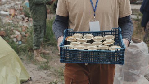 Tracking midsection shot of unrecognizable male social worker or volunteer holding plastic crate with tinned food walking along refugee camp where diverse immigrants living