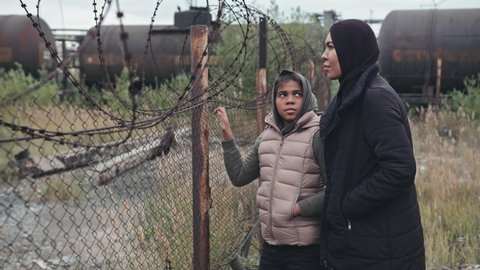 Medium shot of Muslim woman in black hijab and her 11-year-old daughter standing by barbed-wire fence outdoors having conversation