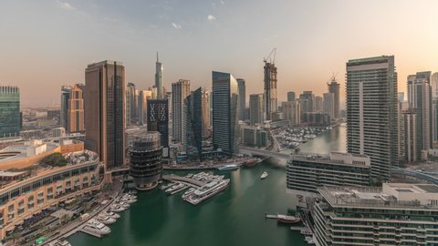 Aerial panoramic view to Dubai marina skyscrapers around canal with floating boats timelapse during sunset with orange sky. White boats are parked in yacht club and near shopping mall
