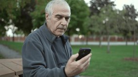 Elderly old man using a smart phone to video chat with family while sitting on a park bench outside