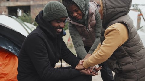 Medium shot of homeless young African-American man in black hoodie playing with two refugee girls sitting outside their tents at immigrants camp