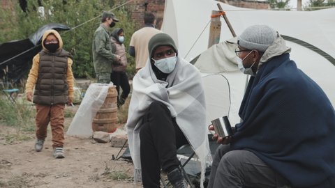 Medium shot of two male immigrants in face masks sitting under blankets outside tents at poor refugee camp having conversation