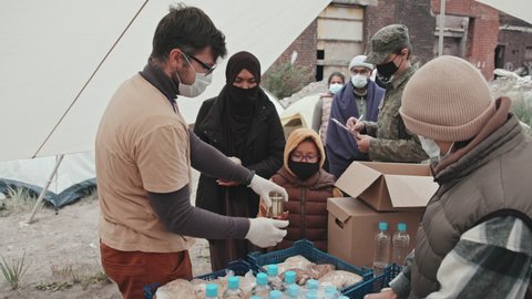 Medium shot of team of volunteers in face masks giving free food to refugees and homeless people living in dirty tent city