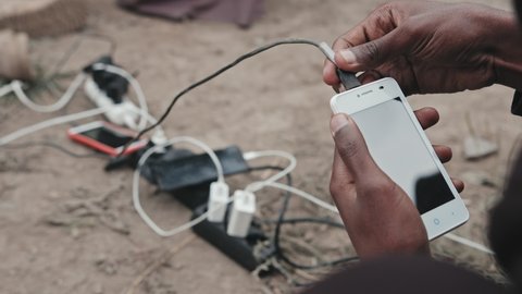 Close-up of unrecognizable African-American refugee man scrolling on smartphone sitting on his laps next to extension cord with chargers