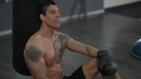 Satisfied tattooed athletic sportsman in boxing gloves sitting at punching bag smiling and looking at camera. Portrait of perspiring confident Middle Eastern man on break training in gym