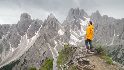 4K  Majestic rocky mountain peaks with woman in foreground wearing yellow jacket and enjoying landscape. Travelling, hiking, backpacking, exploring nature concept. Dolomite Alps, South Tyrol, Italy