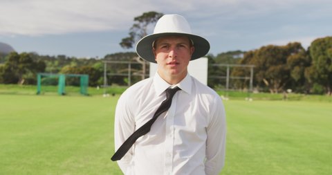 Portrait of a confident Caucasian male cricket umpire wearing white shirt, black tie and a wide brimmed hat, standing on a cricket pitch on a sunny day looking to camera, in slow motion