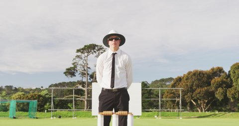 Front view of a Caucasian male cricket umpire wearing white shirt, black tie, sunglasses and a wide brimmed hat, standing on a cricket pitch on a sunny day, gesturing with his hand in slow motion