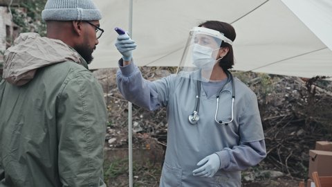 Medium shot of female medical worker in protective clothing, face shield and mask measuring temperature of diverse refugees at tent city