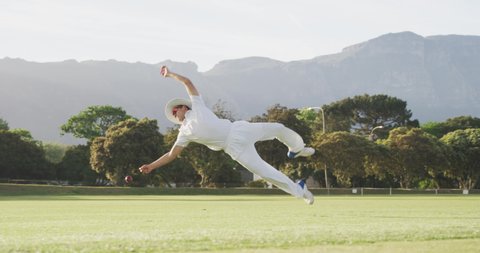 Front view of a Caucasian male cricket player wearing whites and a hat, standing on a cricket pitch, diving for the ball and failing to catch it during a match on a sunny day, in slow motion