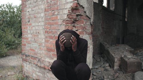 Tilting-down shot of African-American homeless man in Black hoodie sitting on ruins of demolished house on cold day with praying hands